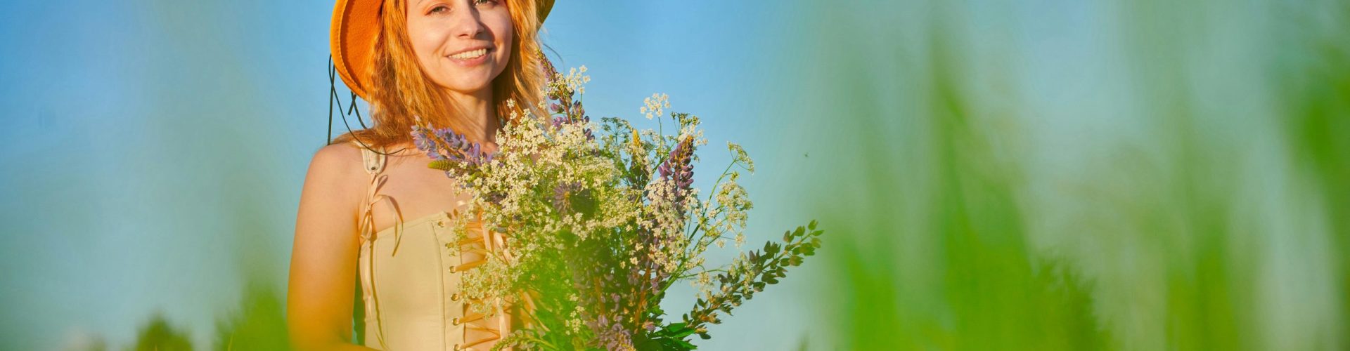 Smiling woman in a field holding a bouquet of wildflowers, wearing an orange hat under a clear blue sky.