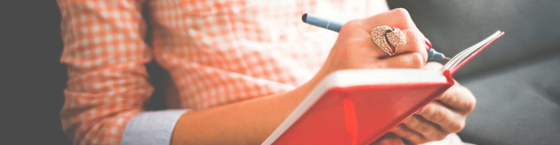 Close-up of a woman writing in a red journal on a sofa, wearing a stylish ring.
