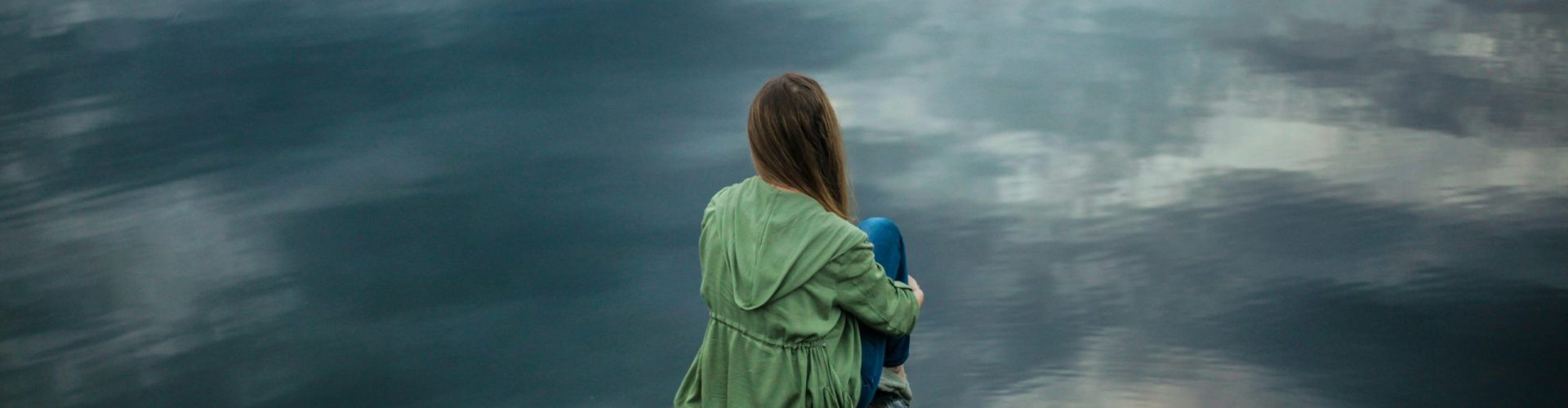 A woman sits on a wooden dock, reflecting by a calm lake under a cloudy sky.