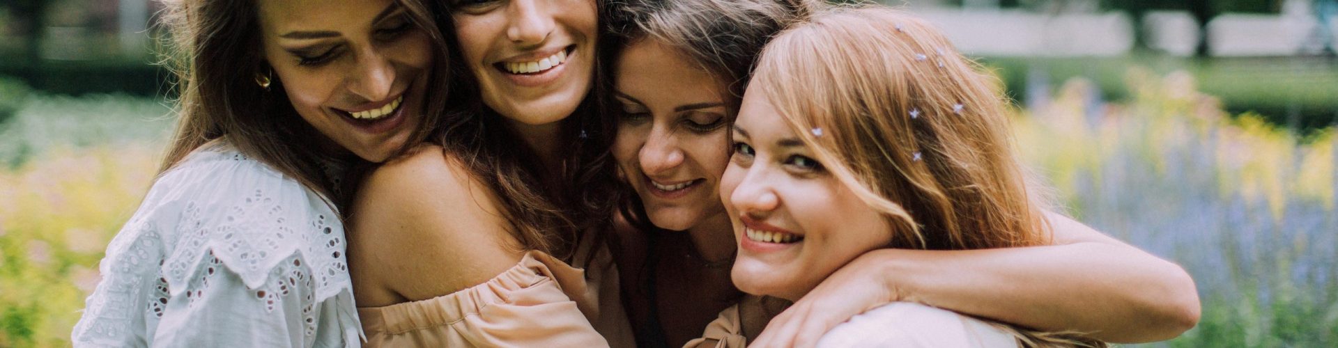 Four smiling women joyfully embrace in a lush green park during summer, showcasing genuine friendship and happiness.