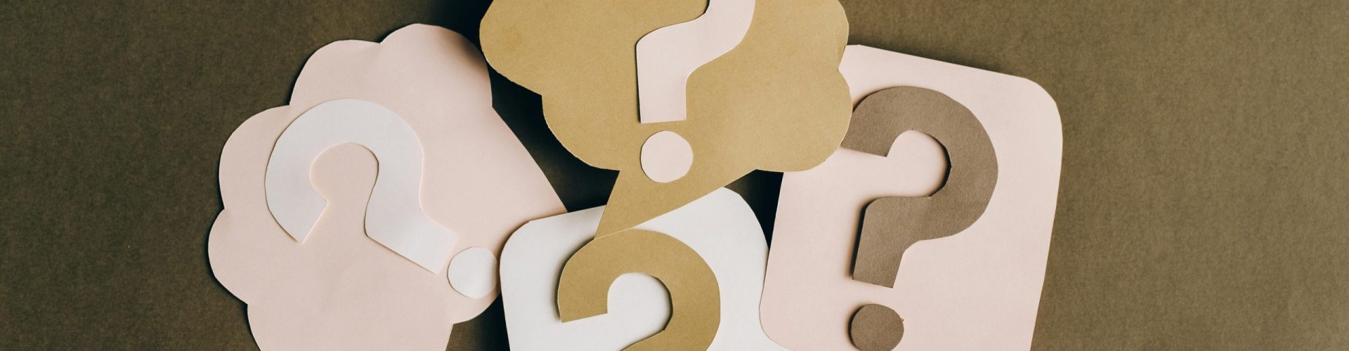 Four paper cutouts of question marks in beige and white on a brown surface, symbolizing inquiry and curiosity.